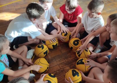 Dzieci w strojach sportowych siedzą w okręgu na podłodze, przed każdym leży żółta piłka.