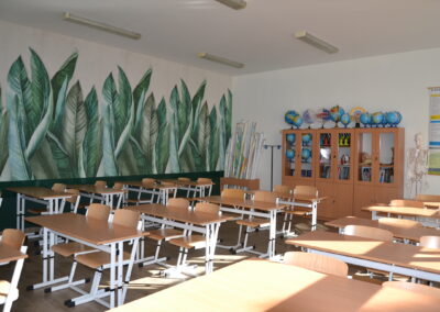 Pusta sala lekcyjna z ławkami szkolnymi ustawionymi w rzędach, na ścianach fototapeta z zielonymi liśćmi.