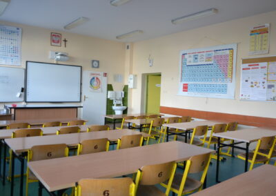 Pusta sala lekcyjna z ławkami szkolnymi ustawionymi w rzędach, na ścianach tablice z układem okresowym pierwiastków.