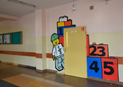 Pusty korytarz szkolny z ilustracją ścienną przedstawiającą figurkę ludzika LEGO i klocki LEGO.